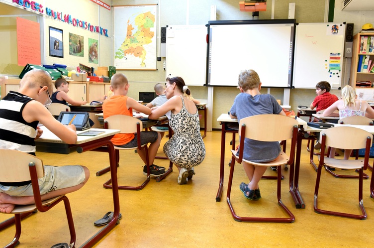 Uczniowie siedzą w ławkach w szkolnej klasie. Nauczycielka kuca przy ławce i rozmawia z uczniem.