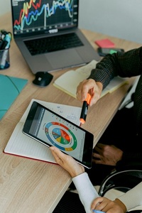 Osoba wskazuje markerem na wykres kołowy na tablecie, obok laptop z wykresami liniowymi