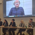 Panel uczestników siedzących przy stole podczas spotkania, z dużym ekranem w tle wyświetlającym transmisję wideo z wystąpienia Pełnomocnika Rządu ds. Osób Niepełnosprawnych Pana Łukasza Krasonia