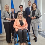 Grupa osób stojących i siedzących przy flagach w sali konferencyjnej, jedna osoba na wózku inwalidzkim na pierwszym planie