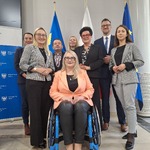 Grupa osób pozująca do zdjęcia przy flagach i roll upie z logo województwa śląskiego w sali konferencyjnej, jedna osoba na wózku inwalidzkim na pierwszym planie