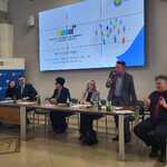 Panel uczestników siedzących przy stole podczas przemówienia Pana Adriana Starońka, Zastępcy Dyrektora Regionalnego Ośrodka Polityki Społecznej Województwa Śląskiego. Duży ekran w tle wyświetlający prezentację o Regionalnym Ośrodku Polityki Społecznej Województwa Śląskiego