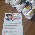 Materiały informacyjne dotyczące spotkania z Powiatowymi Społecznymi Radami ds. Osób Niepełnosprawnych ułożone na stole obok zestawów upominkowych owiniętych w białe materiały z niebieskimi wstążkami i żółtymi długopisami