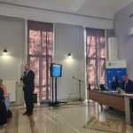 Przewodniczący Sejmiku ds. Osób Niepełnosprawnych Pan Tomasz Jakubiec przy mikrofonie przemawiający podczas spotkania w sali konferencyjnej, obok ekranu z prezentacją i stołu z uczestnikami