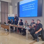 Członek Zarządu Województwa Śląskiego Pan Rafał Adamczyk przemawiający przy stole podczas spotkania w sali konferencyjnej, na ekranie w tle widoczny slajd z napisem: ‘Spotkanie z Powiatowymi Społecznymi Radami ds. Osób Niepełnosprawnych’