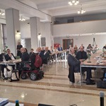 Uczestnicy spotkania siedzący przy stołach w dużej sali konferencyjnej, widoczne osoby korzystające z wózków inwalidzkich i sprzętu wspomagającego