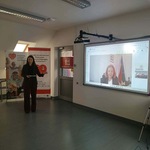 Kobieta stoi w sali szkoleniowej obok ekranu z wideopołączeniem wyświetlanym na projektorze.