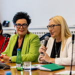 Osoby uczestniczące w spotkaniu siedzą przy stole konferencyjnym. Jedna z osób zabiera głos, trzymając mikrofon w dłoni.