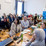 Uczestnicy spotkania siedzą przy stole konferencyjnym i słuchają wystąpienia.