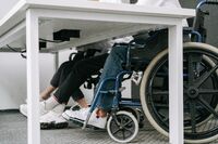 Osoba korzystająca z wózka inwalidzkiego siedzi przy biurku obok drugiej osoby. Na zdjęciu widoczne są dolne części sylwetek, wózek oraz przestrzeń pod blatem stołu, pokazujące dostosowane miejsce do pracy lub spotkania.