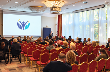 Sala konferencyjna, z uczestnikami konferencji oraz osobą występującą.